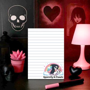 May include: A lined notepad with a cartoon graphic of a grim reaper riding a unicorn on a rainbow, with the text "Apparently im Dramatic". The notepad is on a dark surface, with a pink lamp and other decorative items in the background.