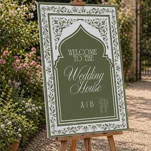 Pode incluir: Um cavalete de madeira exibe uma placa de boas-vindas para um casamento. A placa tem um fundo verde com bordas brancas ornamentadas e o texto "WELCOME TO THE Wedding House". A placa está colocada ao ar livre.