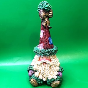 May include: A whimsical Christmas figurine of Santa Claus. The figurine has a long, pointed hat with patches, a white beard, and a smiling face. The hat is topped with a green, textured ornament and a gold bow. The figurine is decorated with green and purple accents.