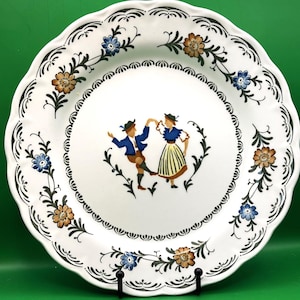 Puede incluir: Plato de cerámica blanca con borde festoneado, decorado con un borde floral negro y una imagen central de dos personas bailando con trajes tradicionales. El plato presenta detalles florales marrones y azules.