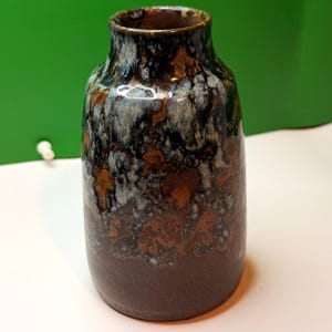 May include: A ceramic vase with a dark brown base and a textured surface. The vase has a speckled pattern of black, white, and bronze colors. The vase has a cylindrical shape with a slightly wider base and a narrow opening at the top.