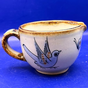 May include: A small, off-white ceramic creamer with a brown rim and handle. The creamer features a blue bird illustration with brown accents and a butterfly design. The creamer has a spout for easy pouring and is suitable for serving cream or milk.