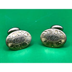 May include: A pair of vintage cufflinks with an oval design. The silver-toned cufflinks feature an engraved floral pattern around the edges and a central monogram. The background is a bright green.