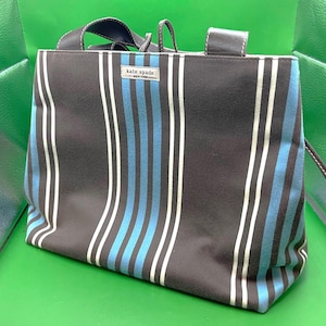 Könnte beinhalten: Eine Tragetasche mit vertikalem Streifenmuster in Schwarz, Weiß und Blau. Die Tasche hat schwarze Griffe und ein kleines Etikett mit der Aufschrift "kate spade NEW YORK". Die Tasche ist aus robustem Material gefertigt.