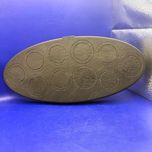 May include: A dark brown, oval-shaped baking mold with ten circular indentations. The mold has a textured surface with a crackle pattern. The background is a solid blue.