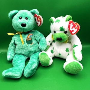 May include: Two plush teddy bears on a green background. One is solid green with a pot of gold and rainbow embroidery. The other is white with green shamrock patterns. Both have red heart-shaped tags and yellow ribbons.