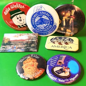 May include: A collection of vintage pinback buttons and a rectangular magnet. The buttons feature various designs, including a lighthouse, fireworks, and a cartoon character. The magnet depicts a covered bridge.