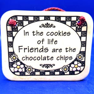 May include: A rectangular ceramic plaque with a white background and a black and white checkered border. The plaque features the quote, "In the cookies of life Friends are the chocolate chips." Decorative flowers and a red rope are also present.