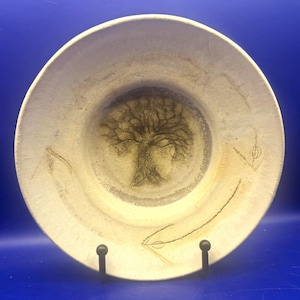 May include: A round ceramic plate with a cream-colored, textured surface. The center features a dark, etched illustration of a tree with intricate branches. Two thin, curved lines resembling arrows or stems are etched along the plate's rim.