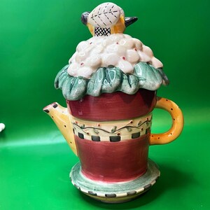 Può includere: Una teiera decorativa in ceramica con coperchio a forma di uccello. La teiera presenta una base rossa, un beccuccio e un manico gialli e un motivo floreale verde e bianco. Il coperchio è sormontato da un uccello giallo e bianco.