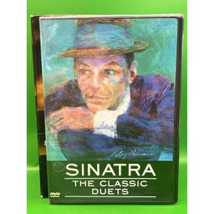 Puede incluir: Caja de DVD con un retrato de Frank Sinatra con sombrero y traje azules. El título "SINATRA" se muestra en letras mayúsculas blancas sobre "THE CLASSIC DUETS" sobre un fondo verde. La caja del DVD es negra.