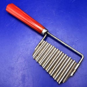 May include: A kitchen crinkle cutter featuring a bright orange, ribbed plastic handle attached to a stainless steel, wavy-edged blade. The metal frame connects the handle to the top of the blade, designed for creating decorative, rippled vegetable cuts.