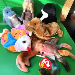 May include: A collection of plush animal toys, including a blue dolphin, a black and white orca, a brown walrus, a colorful fish, a brown crab, and a tan-colored animal. Each toy has a unique design and is made of soft materials.