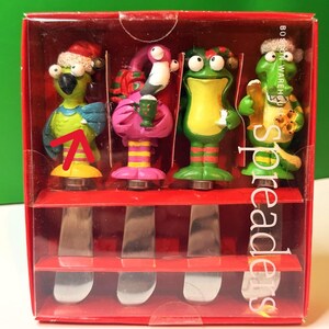 May include: A set of four decorative spreaders in a red box. Each spreader features a whimsical character: a parrot, a flamingo, a frog, and an alligator, all holding drinks. The blades are silver, and the handles are colorful.
