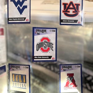 May include: A collection of College Football USA '96 trading cards featuring team logos and names. Cards include West Virginia University, Auburn University, Ohio State University, UCLA Bruins, and University of Alabama Crimson Tide.