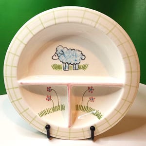 Könnte beinhalten: Eine geteilte Keramikplatte mit einer Schafillustration. Die Platte hat eine cremefarbene Basis mit einem grünen Karomuster. Sie ist in drei Abschnitte mit einem rosa Teiler unterteilt. Das Schaf ist blau mit schwarzen Umrissen, und es gibt rosa Blumen.
