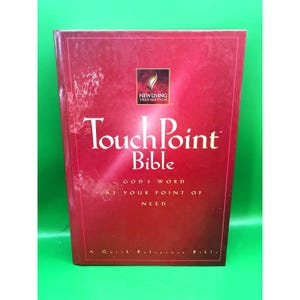Peut inclure: Une Bible "TouchPoint" à couverture rigide rouge avec du texte blanc. La couverture présente le texte "TouchPoint Bible" et "GOD'S WORD AT YOUR POINT OF NEED". Le livre a un logo "New Living Translation".