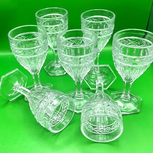May include: A collection of clear, etched glass goblets with hexagonal bases. The goblets feature a detailed floral and geometric pattern, with a slender stem and a wide bowl. The glasses are arranged on a green surface.