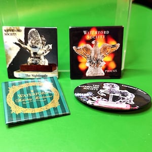 May include: Four Waterford Society collectibles on a green surface. Includes two boxed crystal figurines, one labeled "The Nightingale" and the other "Phoenix." Also, a small book titled "Holiday Heirlooms" and a carousel horse pin.
