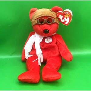 May include: A red plush bear wearing a brown aviator cap with goggles and a white scarf. The bear has a black nose and a circular patch that reads "50 Years of Flight". A heart-shaped tag reads "Ty Beanie Babies".