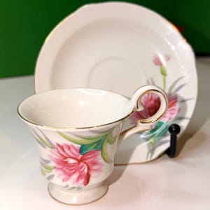 May include: A vintage teacup and saucer set. The cup and saucer are white with a pink floral design and gold trim. The cup has a pedestal base and a curved handle. The saucer has a scalloped edge.
