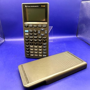Op de afbeelding: Een zwarte Texas Instruments TI-82 grafische rekenmachine met een beschermhoes. De rekenmachine heeft een LCD-scherm en talrijke knoppen voor wiskundige functies. De hoes is lichtgrijs.