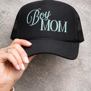 Może przedstawiać: Czarna czapka typu trucker z napisem "Boy Mom" w jasnoniebieskim skrypcie i czcionce blokowej. Czapka ma siatkowy tył i zakrzywiony daszek. Czapka jest trzymana przez dłoń.