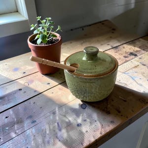 Puede incluir: Un tarro de miel redondo de cerámica verde salvia con tapa a juego y una pequeña muesca para un cazo de madera. El tarro presenta un acabado esmaltado moteado y un pequeño pomo en la tapa. Un cazo de madera de color claro descansa dentro.
