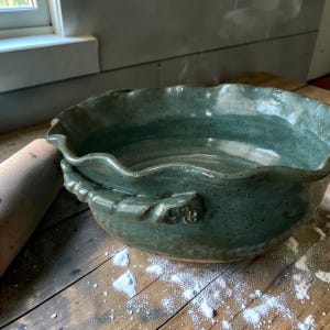 May include: A round, sage green ceramic bowl with a speckled finish, featuring a wavy, scalloped rim and two decorative, twisted-style handles on the sides. The bowl has a glossy glaze and a wide, open basin design for serving or food preparation.