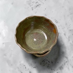 Puede incluir: Un cuenco de cerámica pequeño y redondo con un borde festoneado. El cuenco presenta un esmalte verde salvia moteado con sutiles tonos anaranjados y marrones. La superficie interior muestra estrías concéntricas visibles del torneado.