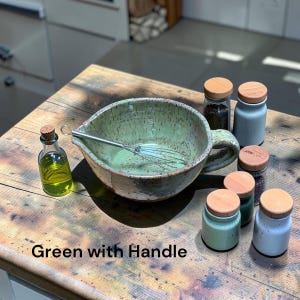 May include: A speckled light green ceramic mixing bowl with a side handle and a metal whisk inside, accompanied by a small glass bottle filled with yellow oil and five small ceramic jars with wooden cork lids. Text reads: Green with Handle.