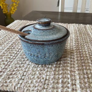 May include: A blue ceramic sugar bowl with a lid and a small wooden spoon. The bowl has a speckled texture and a dark brown rim. The spoon rests in a notch on the side of the lid. The bowl sits on a woven beige placemat.
