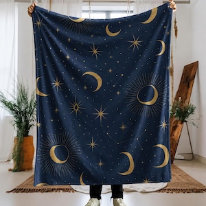 May include: A rectangular throw blanket featuring a deep navy blue background with a repeating pattern of gold celestial elements, including various phases of the moon, suns with radiating lines, and multi-pointed stars of different sizes.