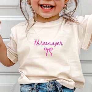May include: Cream-colored t-shirt with the word "threenager" in pink script and a pink bow graphic. The shirt is made of soft material and is suitable for toddlers. The shirt is paired with blue denim jeans.