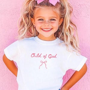 May include: A young girl with blonde hair and a pink bow smiles, wearing a white t-shirt with the text "Child of God" in pink. The background is a solid pink wall.