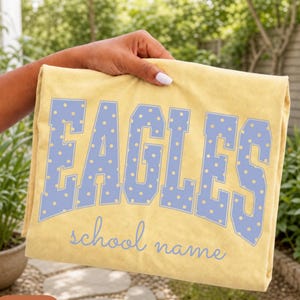 May include: A butter yellow t-shirt featuring the word EAGLES in large, light blue block letters with a yellow polka dot pattern. Below the word EAGLES, the phrase school name is printed in a light blue cursive script font on the fabric.