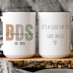 May include: White ceramic mug with black handle and rim. One side features the text "DDS Jasmine EST. 2026" in brown, gray, and green. The other side reads "IT'S A GOOD DAY TO SAVE SMILES" with a heart.
