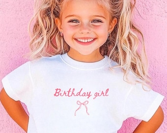 May include: A young girl with blonde hair and a pink bow smiles, wearing a white t-shirt with the words "Birthday girl" in pink script and a bow graphic. She is standing in front of a pink wall.