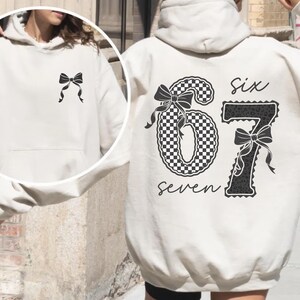 May include: Sand-colored hoodie with a black bow on the front and a large graphic on the back. The back graphic features the numbers "67" with a checkered pattern, bows, and the words "six" and "seven".