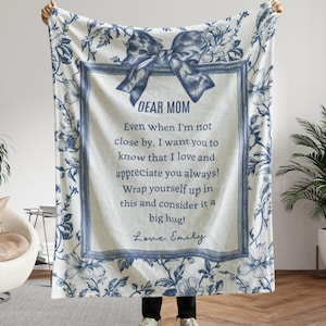 Pode incluir: Um cobertor branco com um padr&atilde;o floral azul e uma mensagem central emoldurada que diz "DEAR MOM". A mensagem inclui uma nota pessoal e &eacute; assinada "Love, Emily". Uma grande fita azul est&aacute; no topo.