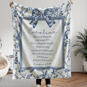 May include: A white and blue floral throw blanket with a bow design and the name "Caroline" in script. The blanket features a framed text with a religious quote from Numbers 6:24-26. The blanket is a soft, cozy home decor item.
