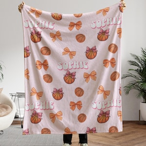 May include: A light pink blanket featuring a repeating pattern of basketballs, bows, and the name "Sophie" in a bubble font. The basketballs have pink bows. The bows are orange. The blanket is held up by a person.