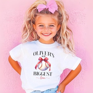 Puede incluir: Una camiseta blanca con el texto "OLIVER'S BIGGEST fan" y un gr&aacute;fico de b&eacute;isbol con una cinta roja. La camiseta la lleva una ni&ntilde;a con cabello rubio y un lazo rosa, sonriendo frente a un fondo rosa.