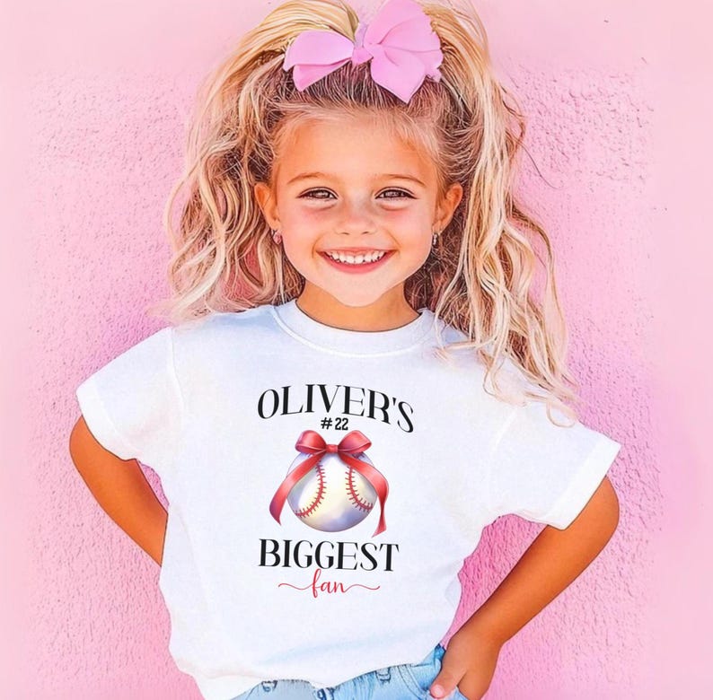 Puede incluir: Una camiseta blanca con el texto "OLIVER'S BIGGEST fan" y un gr&aacute;fico de b&eacute;isbol con una cinta roja. La camiseta la lleva una ni&ntilde;a con cabello rubio y un lazo rosa, sonriendo frente a un fondo rosa.