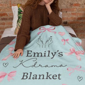 May include: A light blue blanket with pink bows, hearts, and the text "Emily's Kdrama Blanket." The blanket features hand-drawn illustrations and is designed for comfort and warmth. A cozy home decor item.