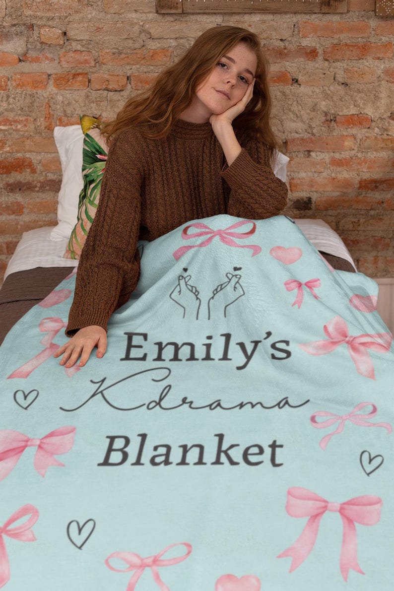 May include: A light blue blanket with pink bows, hearts, and the text "Emily's Kdrama Blanket." The blanket features hand-drawn illustrations and is designed for comfort and warmth. A cozy home decor item.