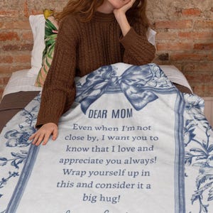 Pode incluir: Um cobertor branco com um design floral azul e o texto "DEAR MOM" em uma moldura decorativa. O cobertor inclui uma mensagem de amor e apre&ccedil;o, assinado "Love, Emily".