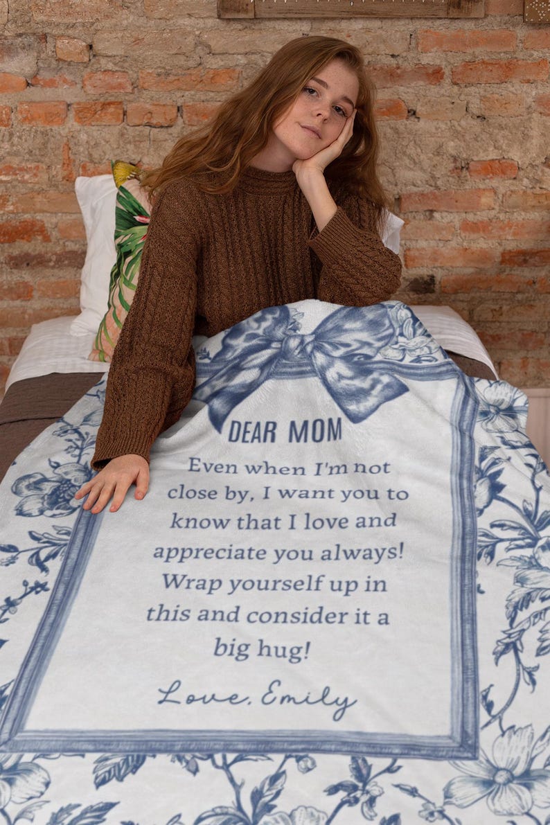 Pode incluir: Um cobertor branco com um design floral azul e o texto "DEAR MOM" em uma moldura decorativa. O cobertor inclui uma mensagem de amor e apre&ccedil;o, assinado "Love, Emily".