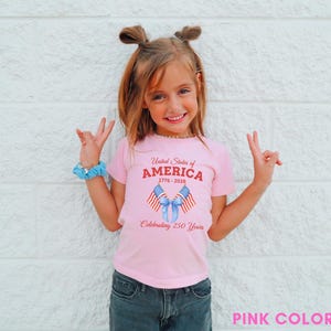 May include: A pink t-shirt with the text "United States of America 1776-2026 Celebrating 250 Years" and a graphic of two American flags tied with a blue ribbon. The shirt is worn by a child.