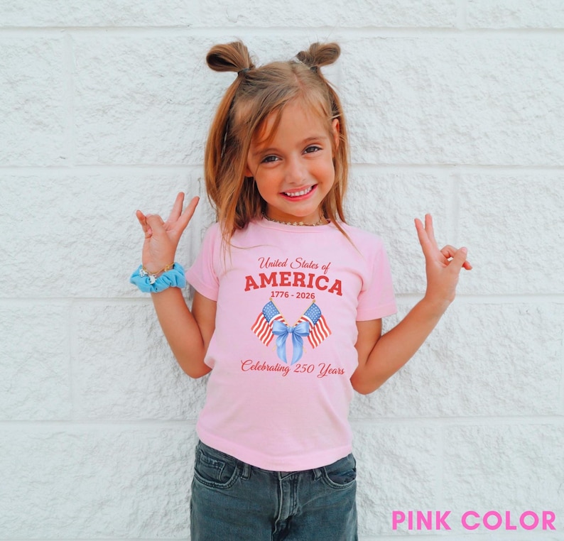 May include: A pink t-shirt with the text "United States of America 1776-2026 Celebrating 250 Years" and a graphic of two American flags tied with a blue ribbon. The shirt is worn by a child.